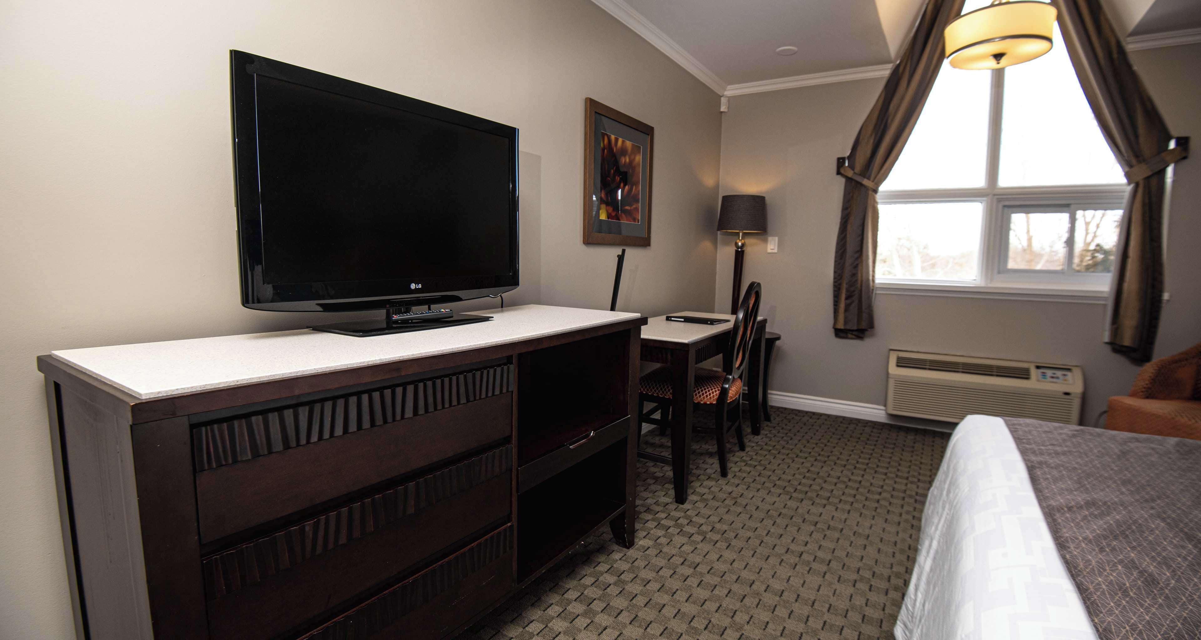 Habitación de hotel con un televisor de pantalla plana grande sobre un mueble blanco, un escritorio con una silla, una cama con colcha de diseño gris y blanco, una ventana con cortinas y una lámpara fijada a la pared.