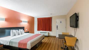A hotel room with a bed, a desk, a chair, a television, and an air conditioning unit.