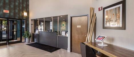 A hotel reception area with a herringbone wood floor, a reception desk with a digital screen, and a framed artwork on the wall.