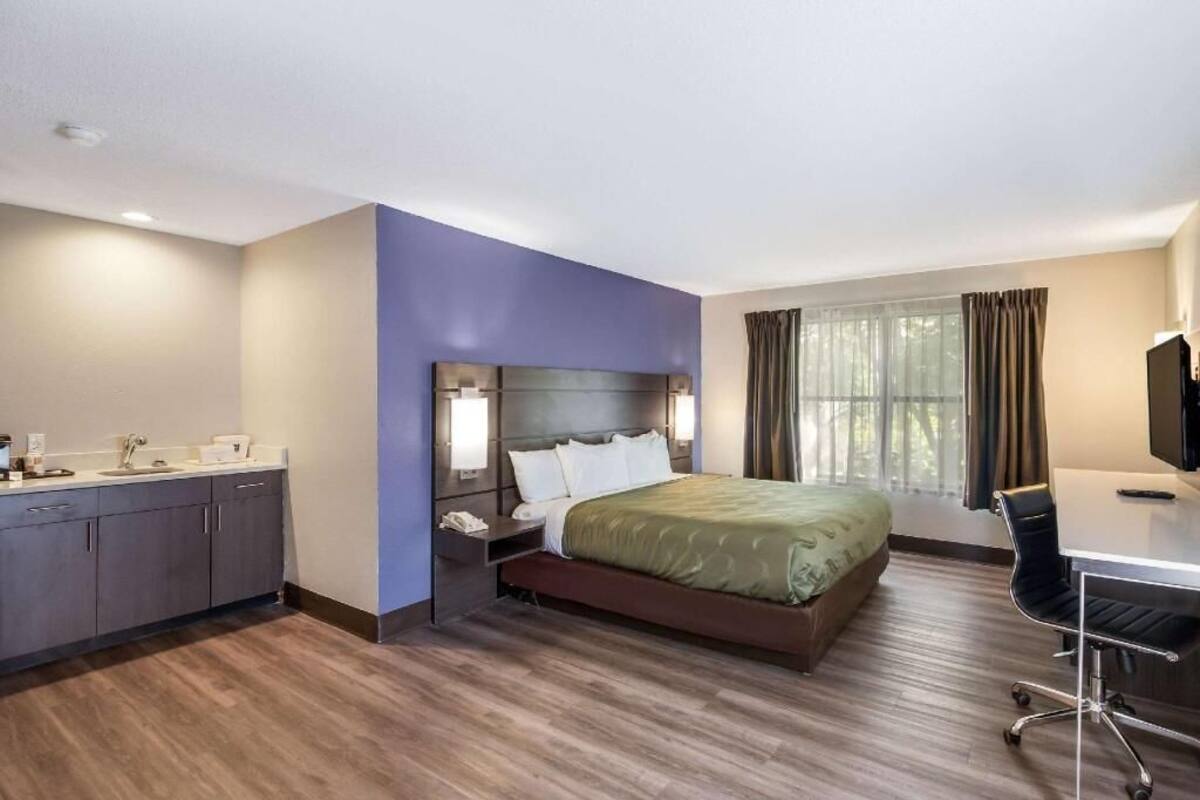 Une chambre d’hôtel comprenant un lit, un bureau, une chaise, une table de chevet, une lampe, une fenêtre avec des rideaux et une salle de bain avec un lavabo et un miroir.