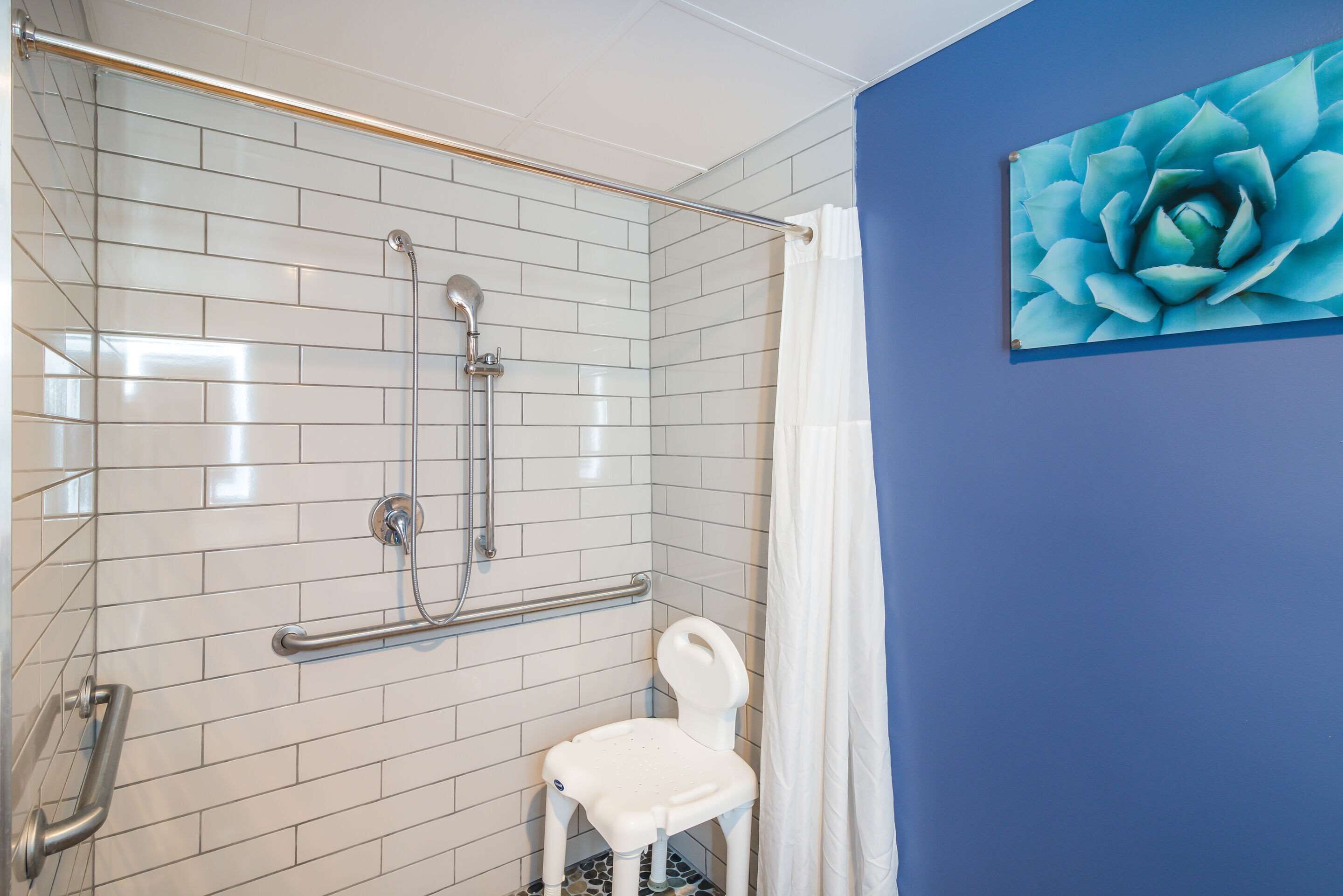 A bathroom with white tiled walls, a shower with a glass door, a white chair, and a blue wall with a large flower painting.