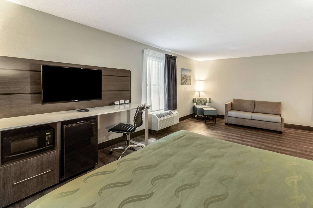 Une chambre d’hôtel comprenant un lit, une télévision, un bureau avec un ordinateur, une chaise, un canapé et une fenêtre avec des rideaux.
