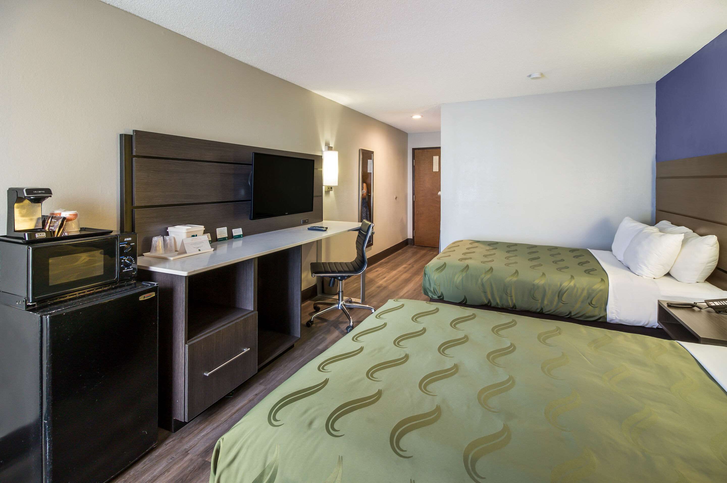 A hotel room with a kitchenette, a bed with green patterned bedding, a desk with a flat-screen TV, and a chair.