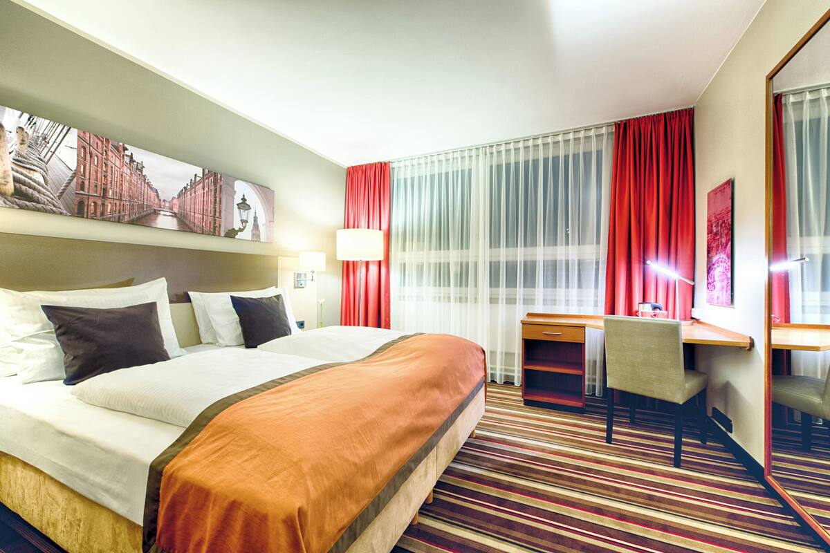 A hotel room with a large bed, a desk, and a chair. There is a window with curtains and a painting on the wall.