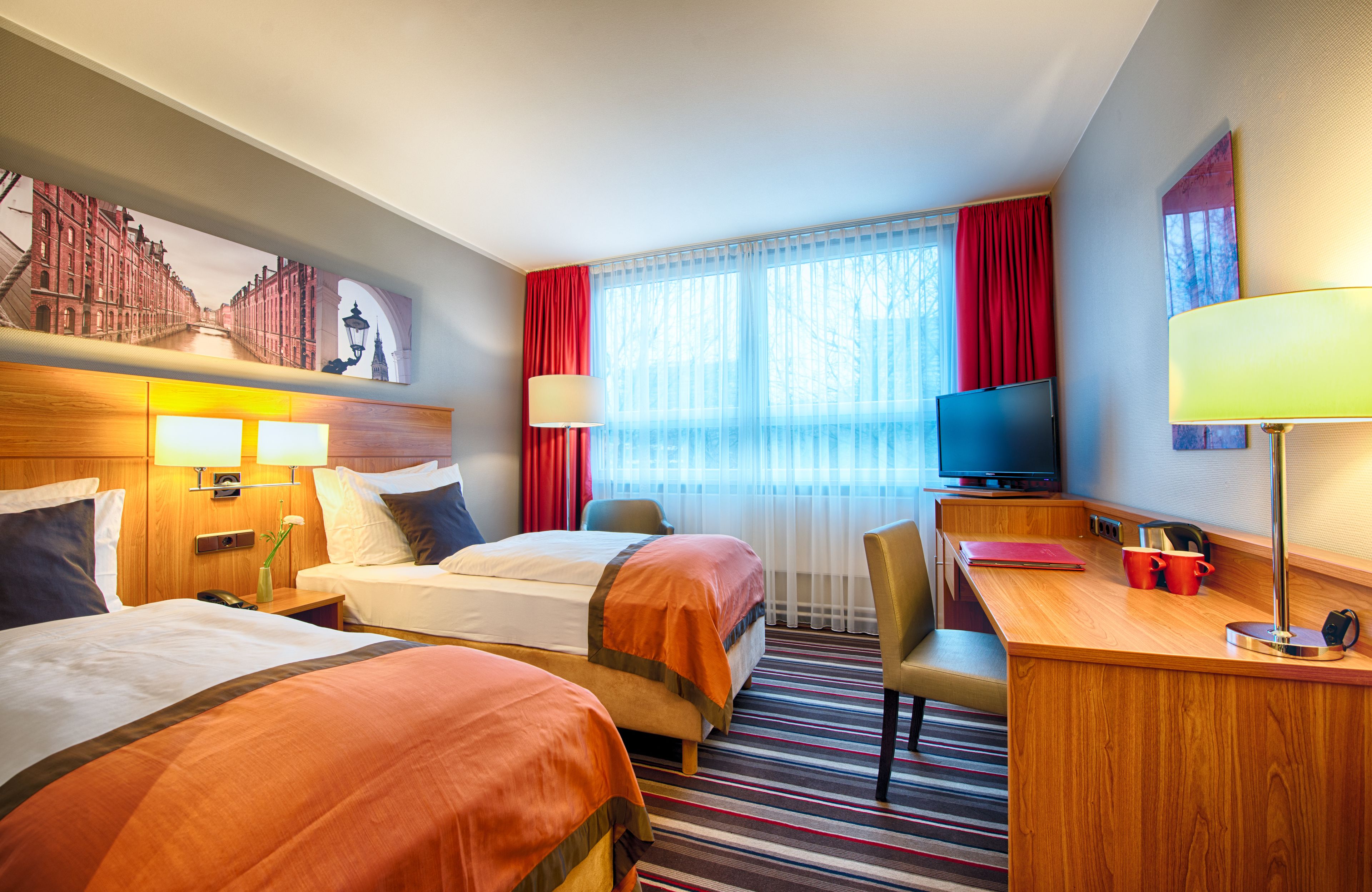 Habitación de hotel con dos camas, un escritorio, un televisor y un ventanal con cortinas.