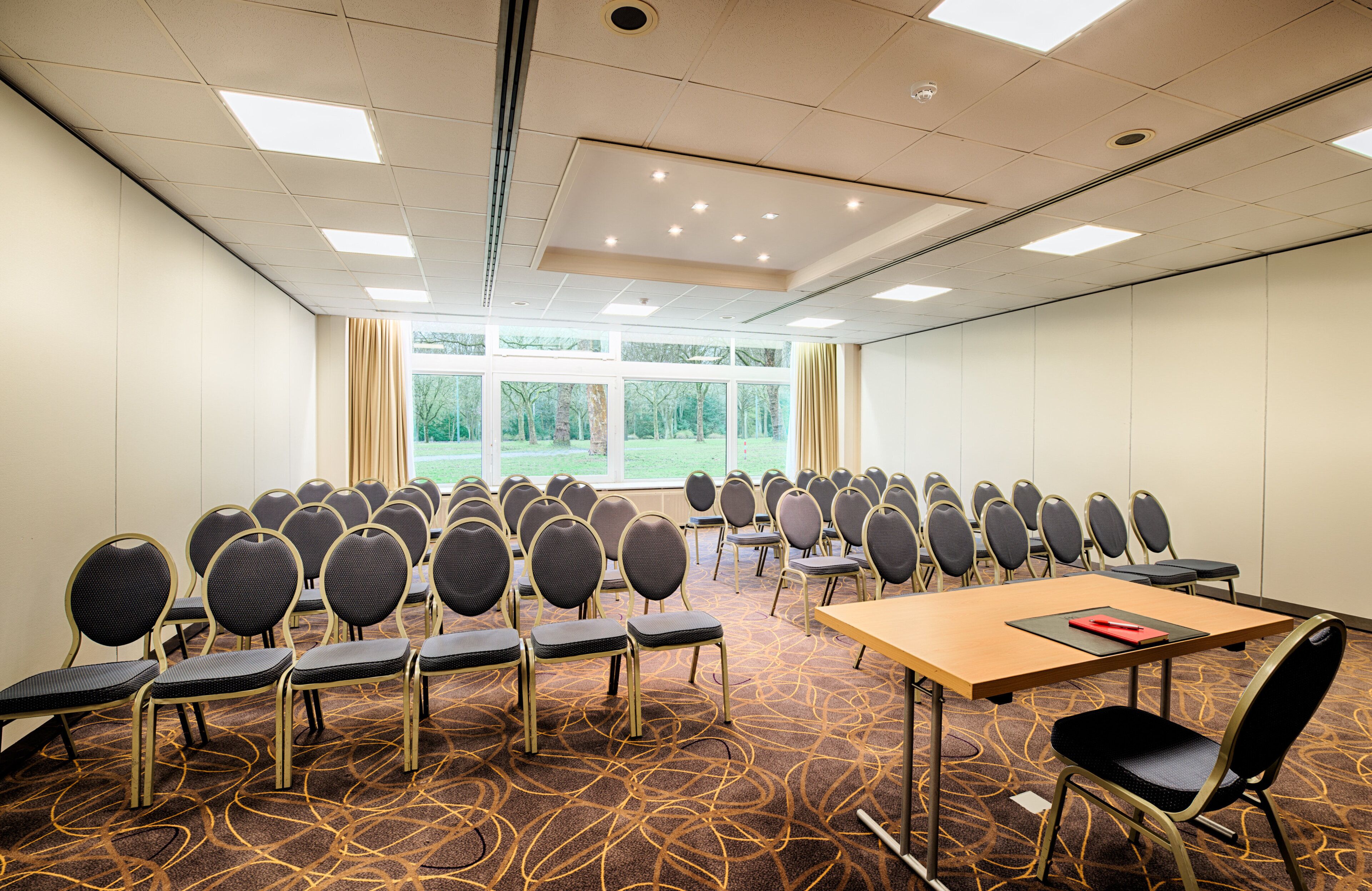 Un salón de conferencias con filas de sillas, una mesa con una carpeta roja y ventanas grandes con vistas a árboles.