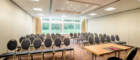 A conference room with rows of chairs, a table with a red folder, and large windows with a view of trees.