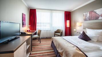 A hotel room with a bed, a desk with a television, a chair, a window with curtains, and a painting on the wall.