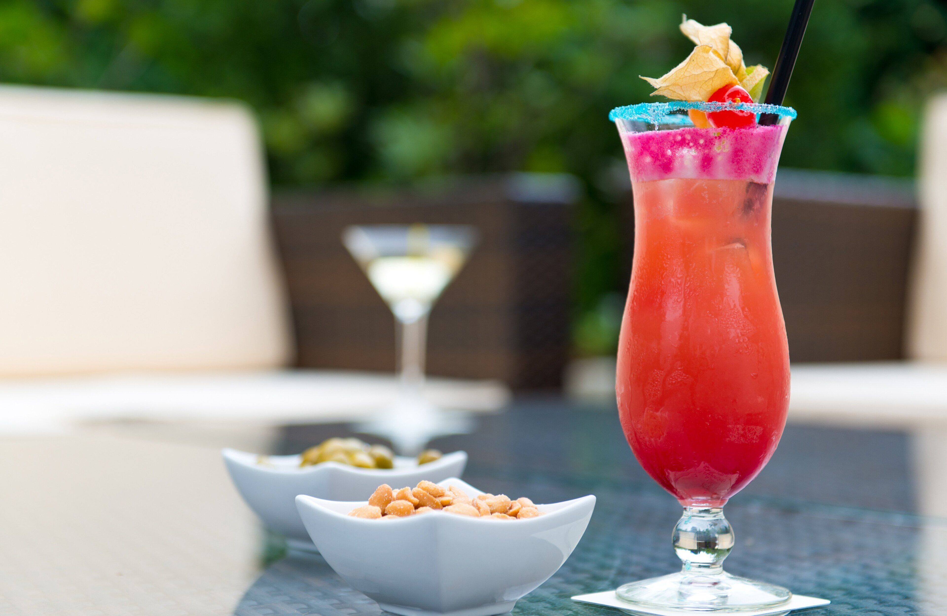 Een cocktailglas met een roze drankje, versierd met fruit en een suikerrand, op een tafel met een onscherpe buitenachtergrond.
