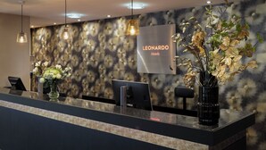 En hotellreception med modern design, med en vägg med ett mönster, en skylt med texten ”Leonardo Hotels” och två vaser med blommor.