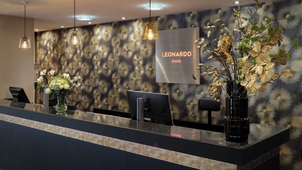Un taulell de recepció d'hotel de disseny modern, amb una paret amb dibuixos, un cartell de 'Leonardo Hotels' i dos vasi amb flors.