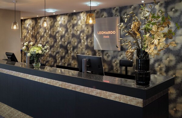Un mostrador de recepción de hotel con diseño moderno, que presenta una pared estampada, un letrero que dice 'Leonardo Hotels' y dos jarrones con flores.
