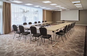 Una sala de conferencias con una larga mesa, sillas, una pantalla de proyector y un amplio ventanal con cortinas.