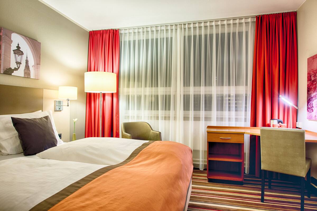 A hotel room with a bed, desk, chair, and curtains.