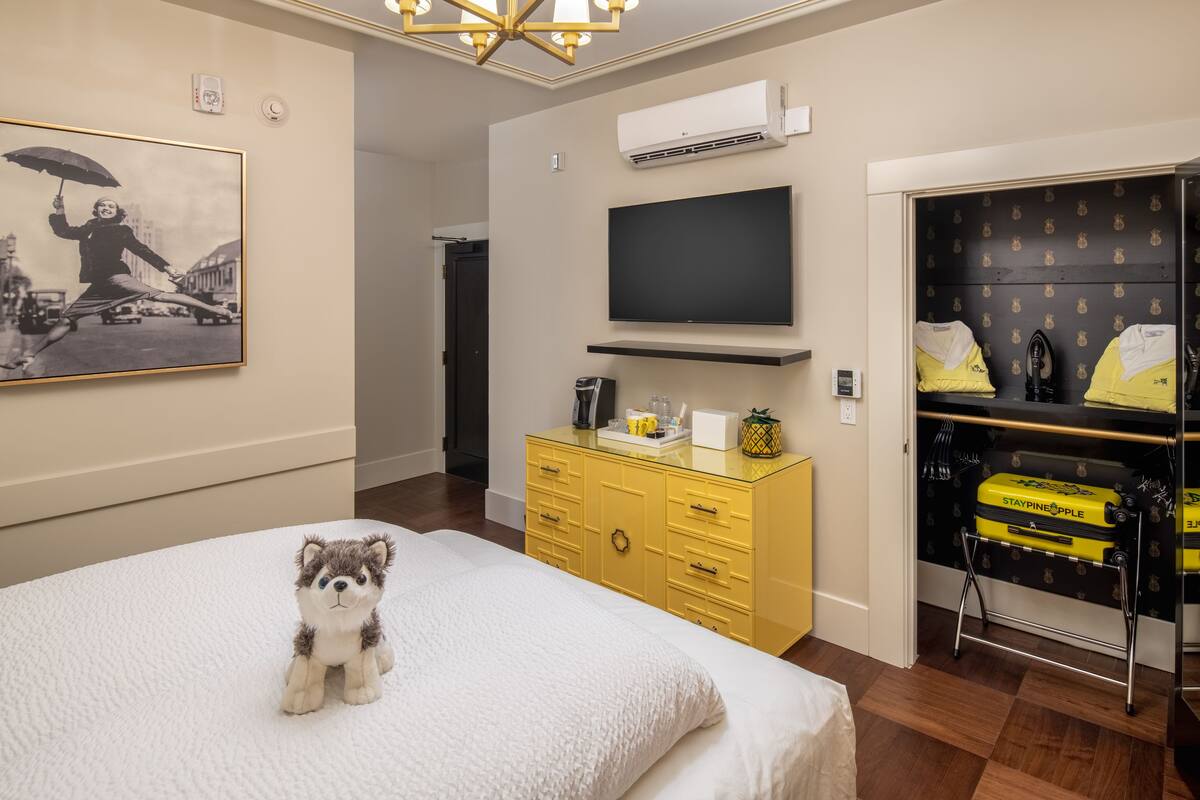 A hotel room with a bed, a TV mounted on the wall, a yellow dresser, and a closet with luggage.