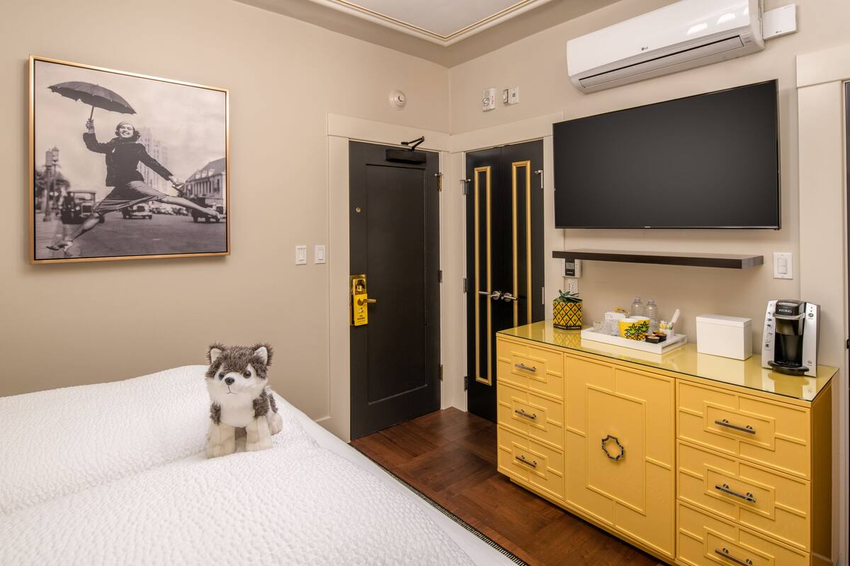 A hotel room with a bed, a yellow dresser, a flat-screen TV, and a framed photo on the wall.