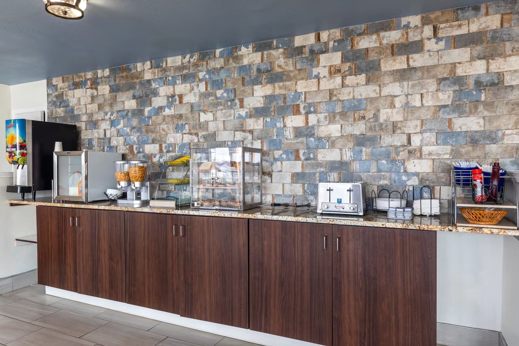 A kitchen with a brick backsplash, a refrigerator, a coffee machine, a toaster, and a juice dispenser.