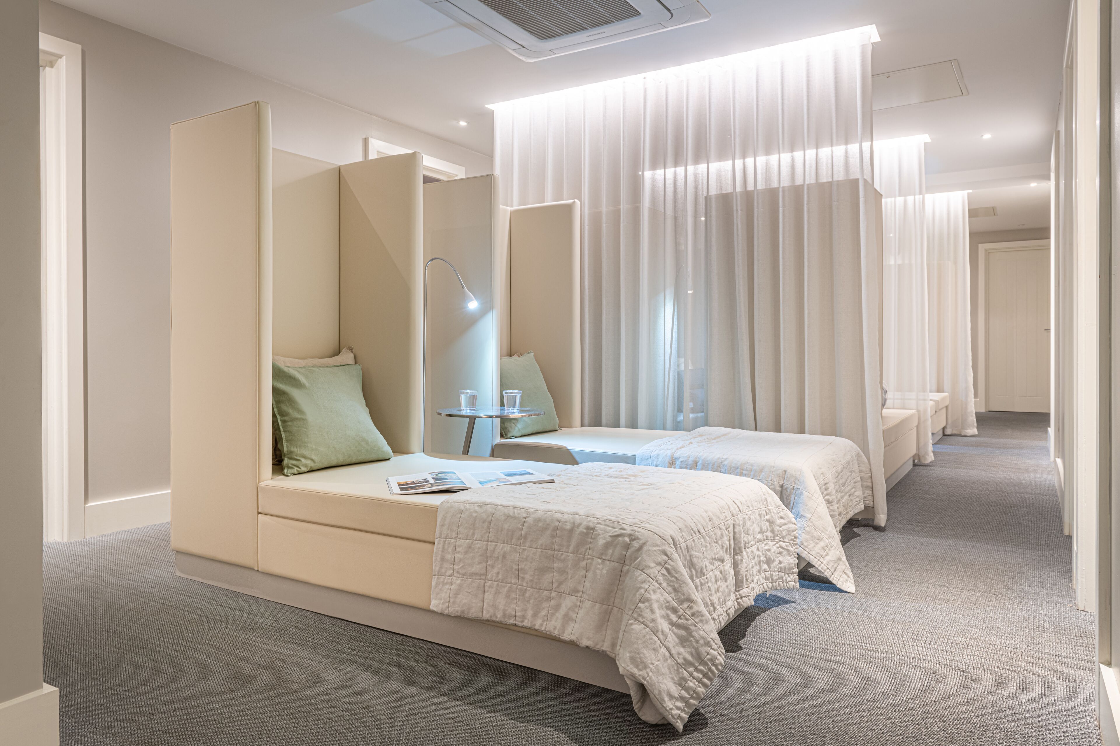 A hotel room with two beds, a bedside table with a lamp, and a large window with sheer curtains.