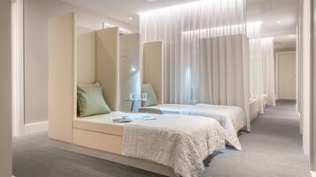 A hotel room with two beds, a bedside table with a lamp, and a large window with sheer curtains.