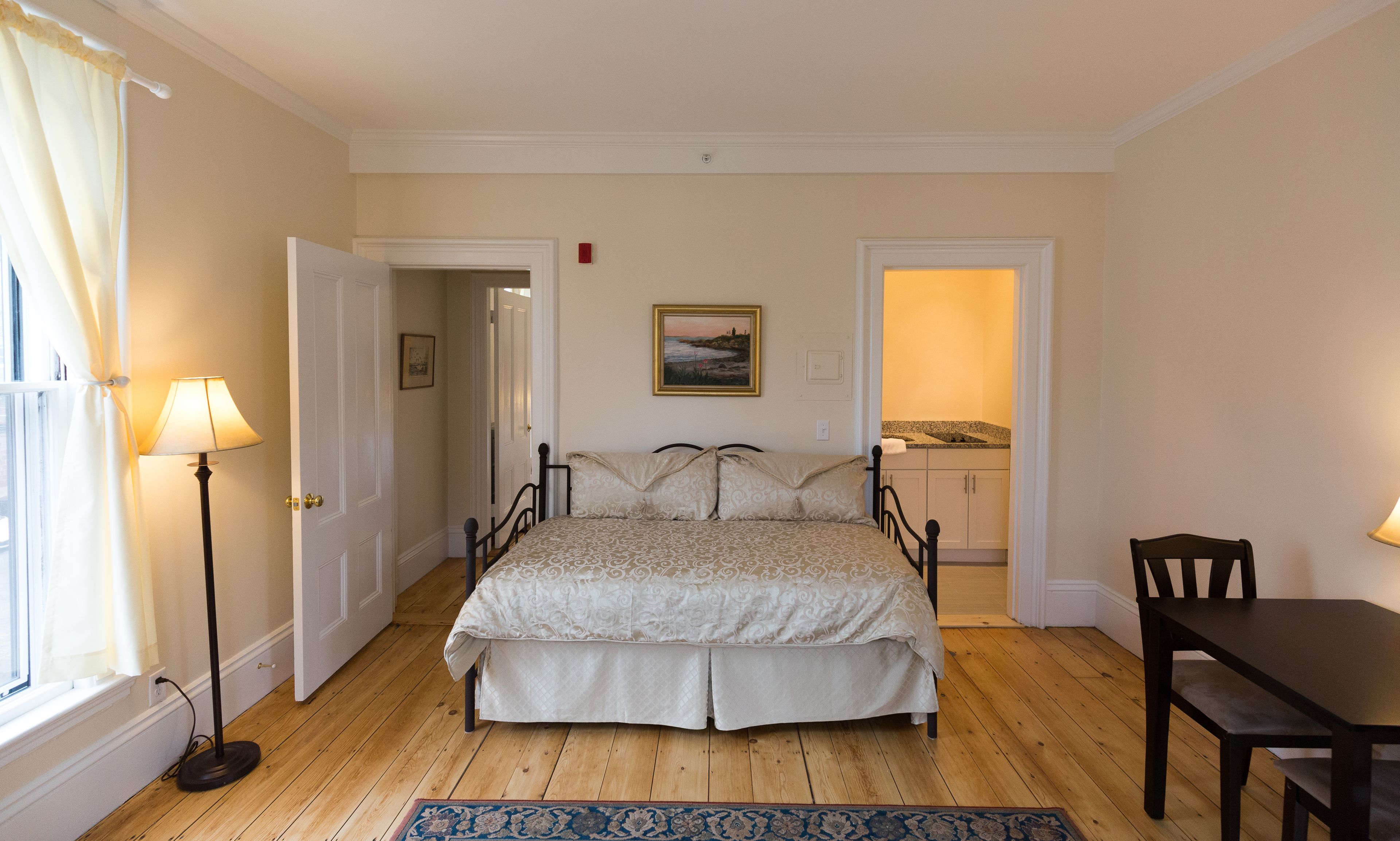 A bedroom with a bed, a bedside table, a chair, a lamp, a painting, and a view of the bathroom.