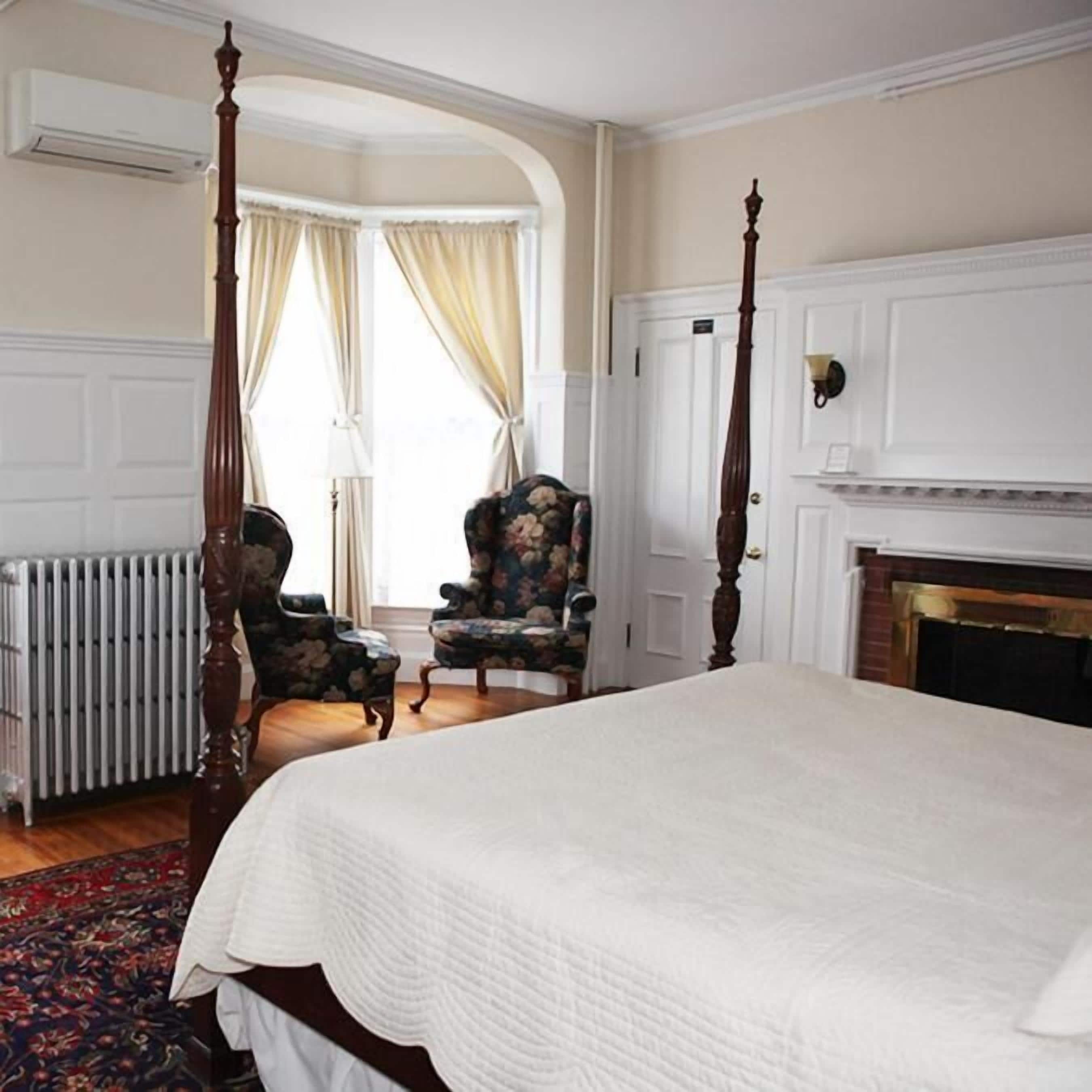 A four-poster bed with a white bedspread, two upholstered chairs with floral upholstery, a white radiator, and a fireplace.