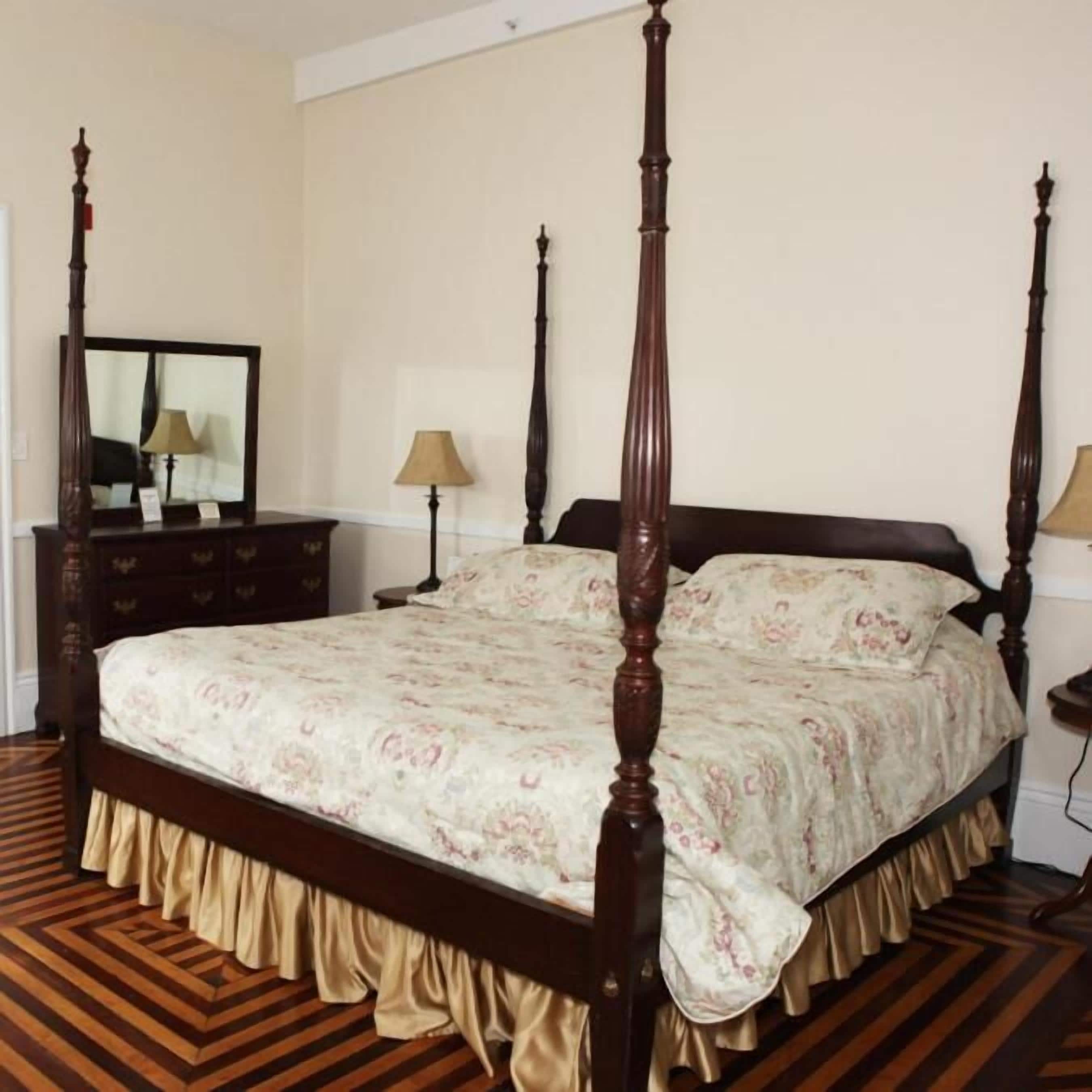 Una cama con dosel adornada con colcha floral y falda con volantes, ubicada en una habitación con piso de parquet de madera y cómoda con espejo.