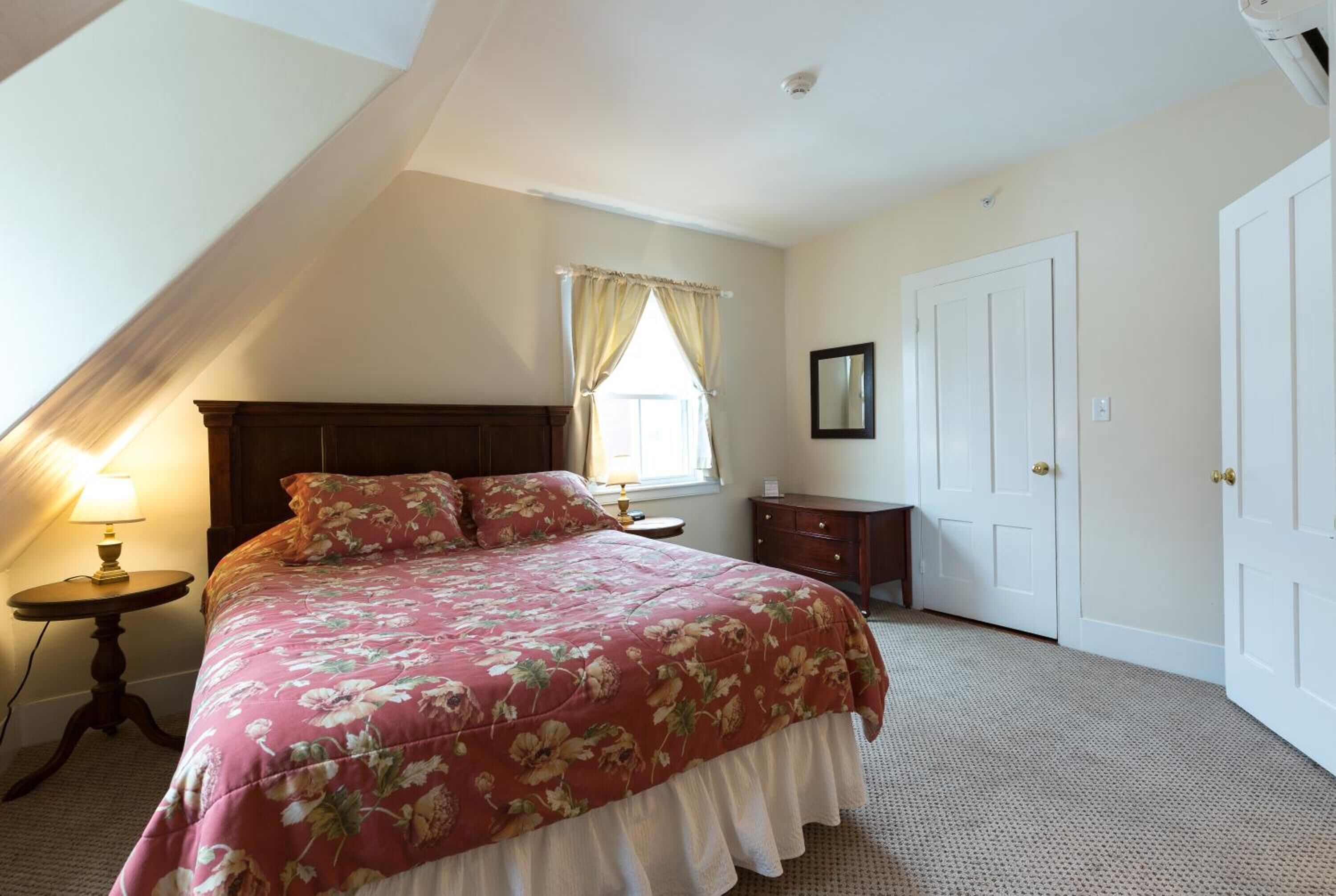 A bedroom with a bed, a nightstand, a lamp, a window with curtains, a wardrobe, and a mirror.