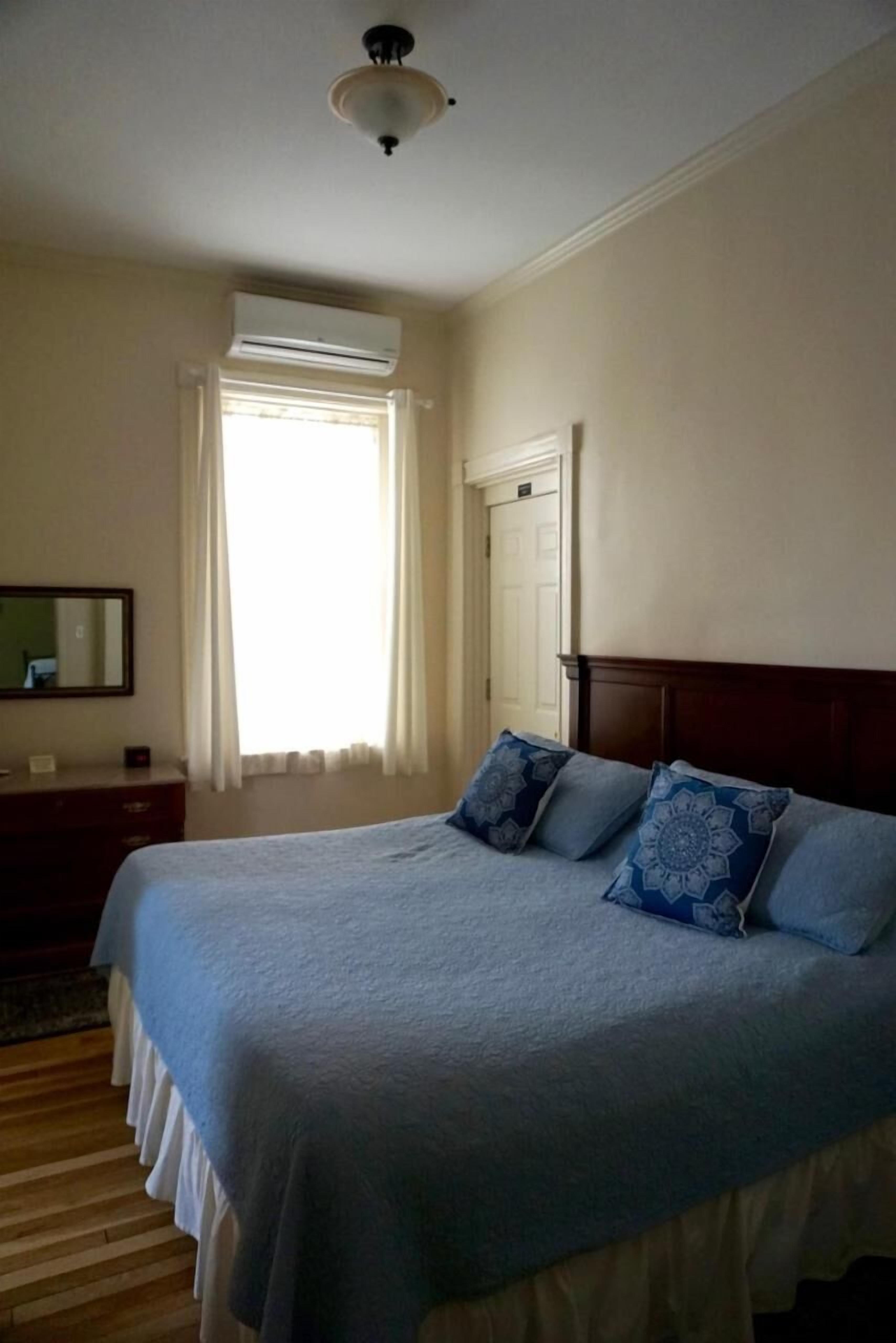 Une chambre à coucher comprenant un lit, une commode, un appareil de climatisation, une fenêtre avec des rideaux et une porte.