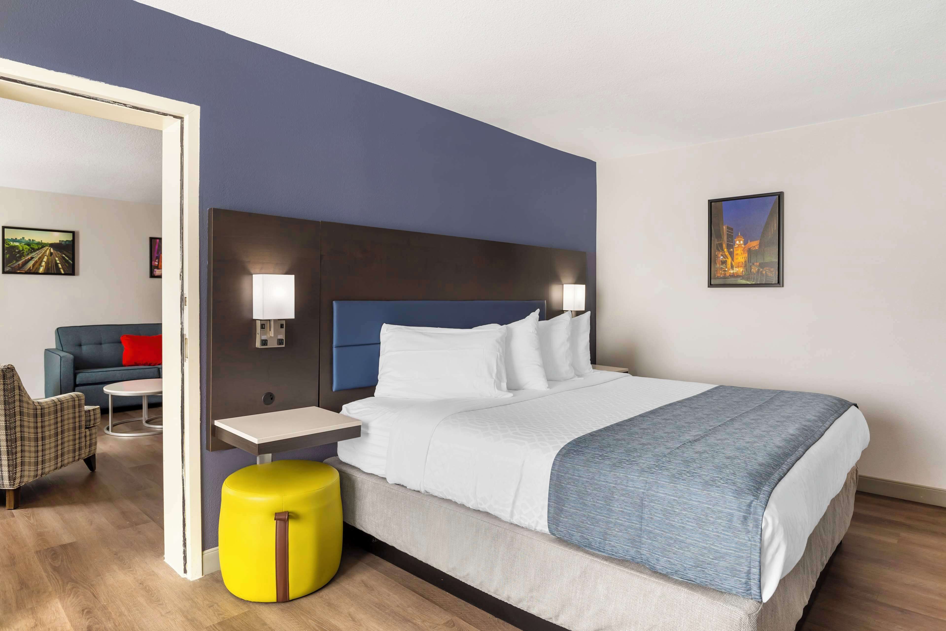 A hotel room with a bed, a nightstand, a chair, a small table, and a yellow ottoman.