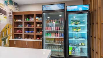 A hotel lobby with wooden cabinets stocked with snacks and beverages, a refrigerator with a variety of drinks, and a wall mural depicting a historic building.