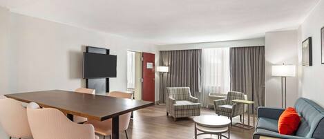 A modern hotel room with a wooden dining table, two pink chairs, a blue sofa with a red pillow, a plaid armchair, and a small round table.