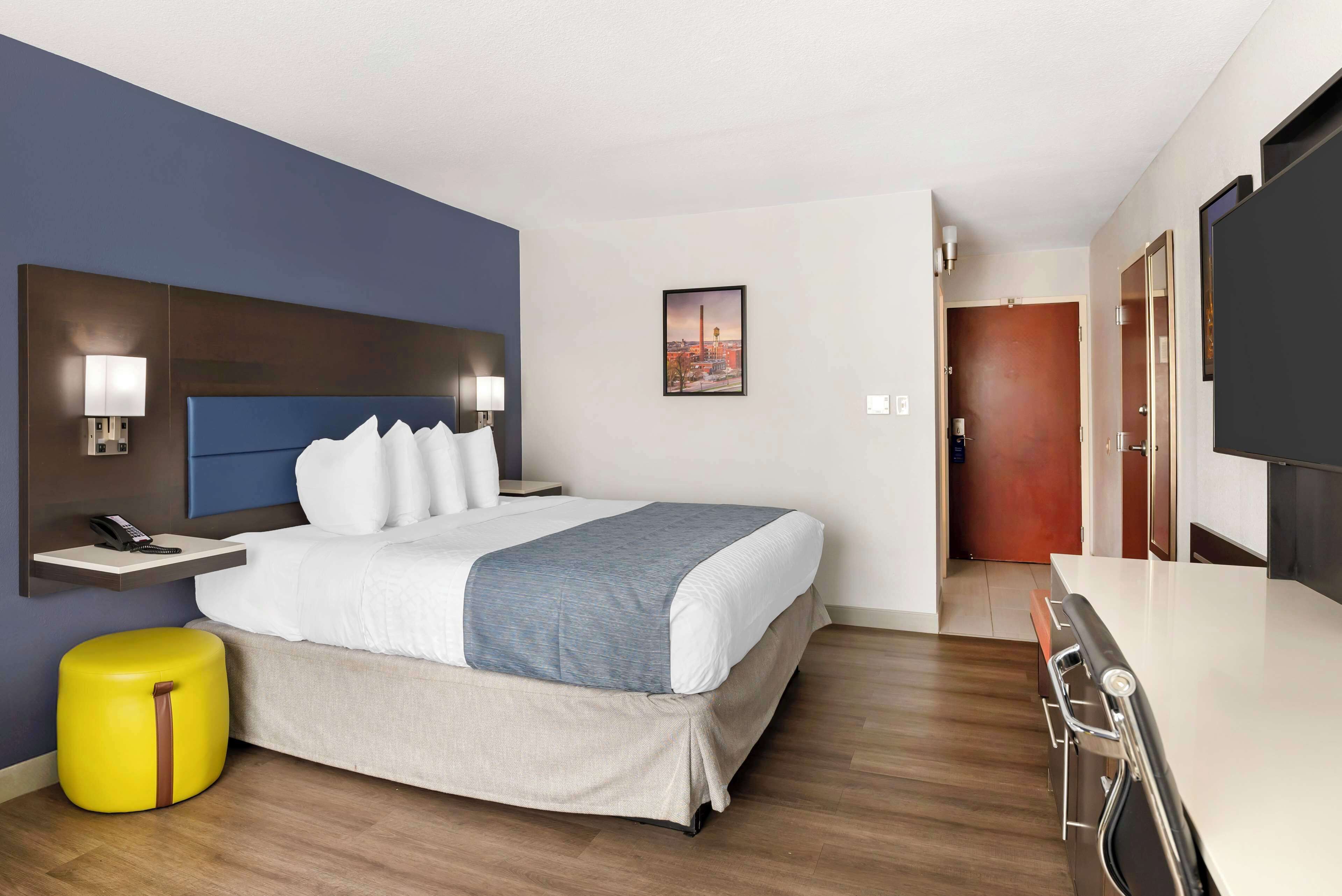 Una habitación de hotel con cama, mesita de noche, lámpara y un pequeño otoman amarillo.
