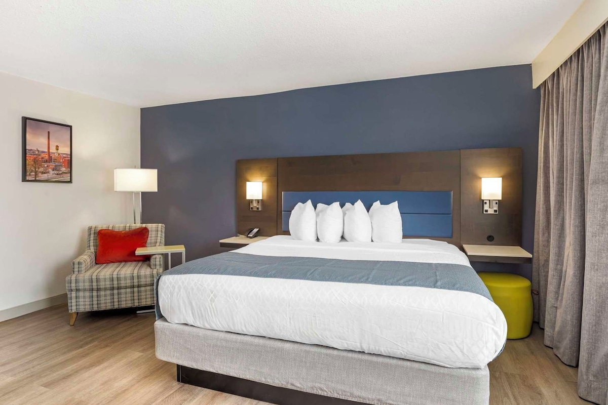 Habitación de hotel con una cama grande, una mesita de noche, una silla, una lámpara y un cuadro en la pared.