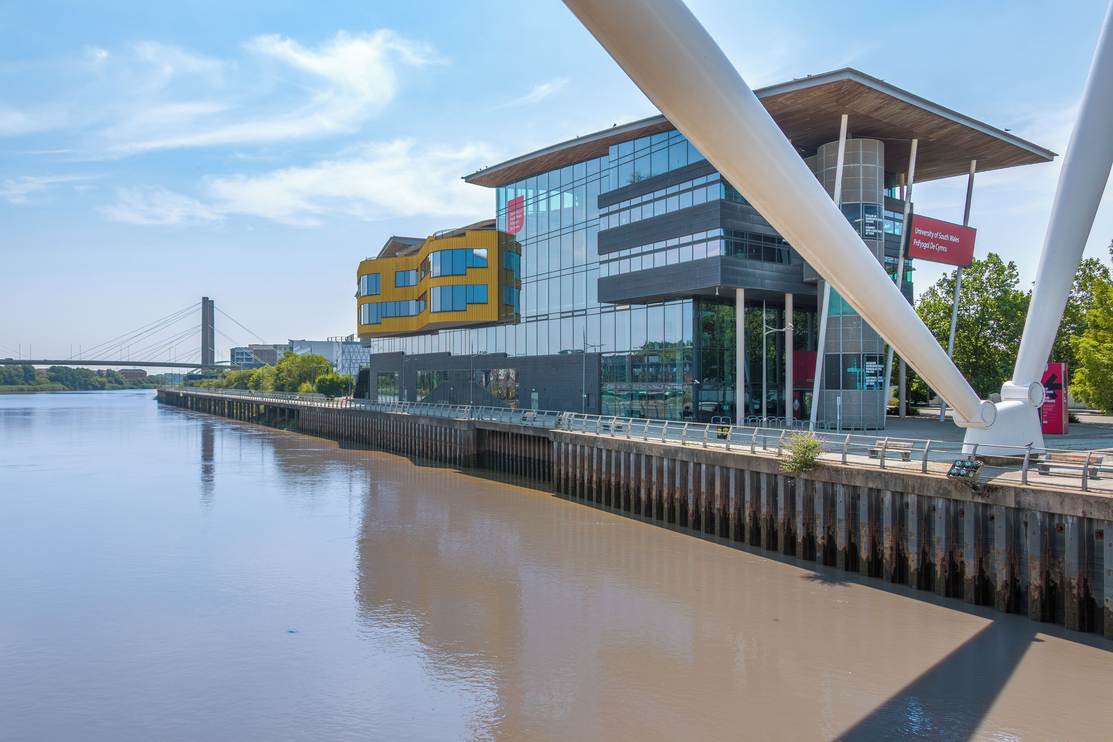A modern building with a glass facade, a wooden structure on the right, and a river in the foreground.