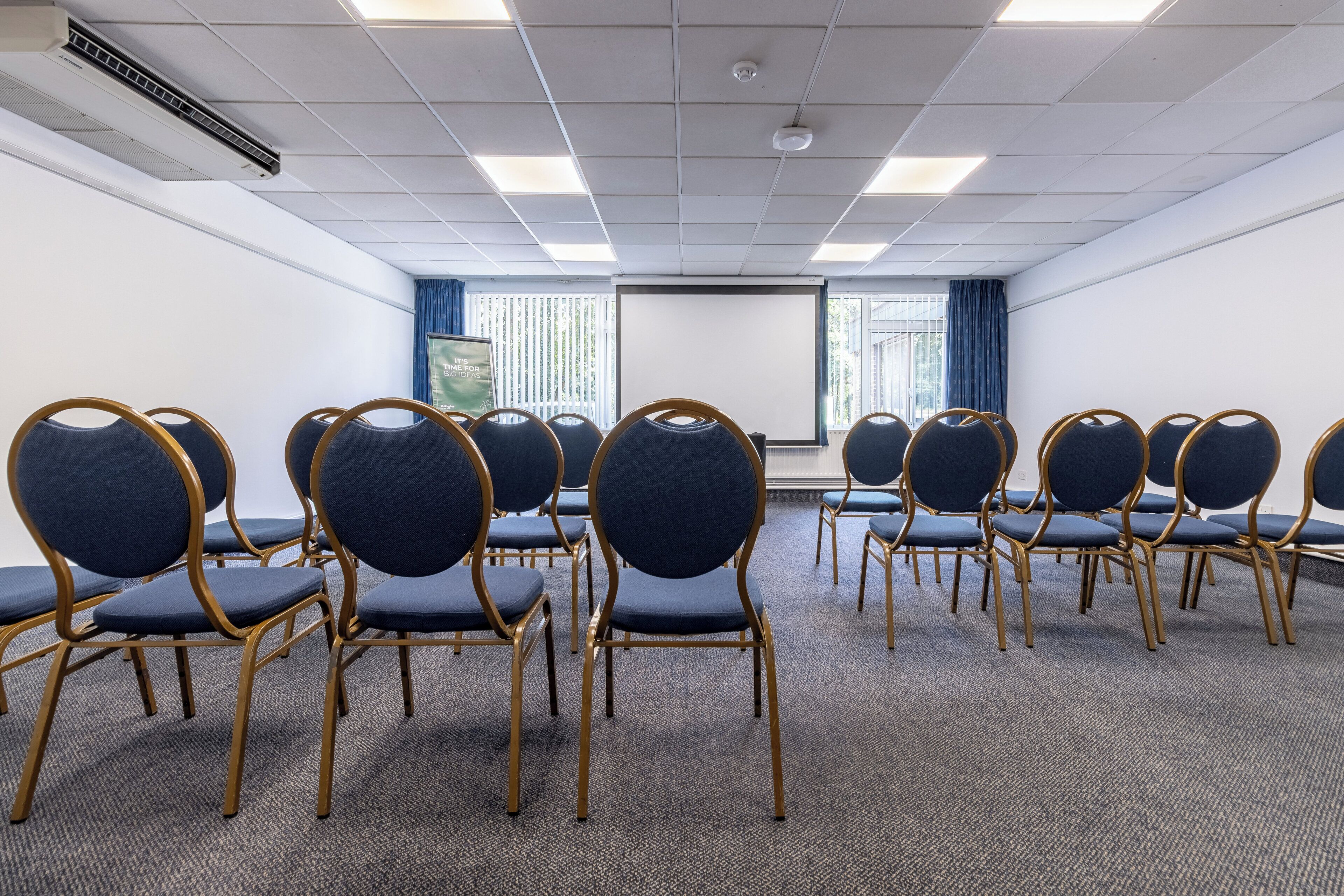 A conference room with rows of chairs, a projector screen, and windows with curtains.