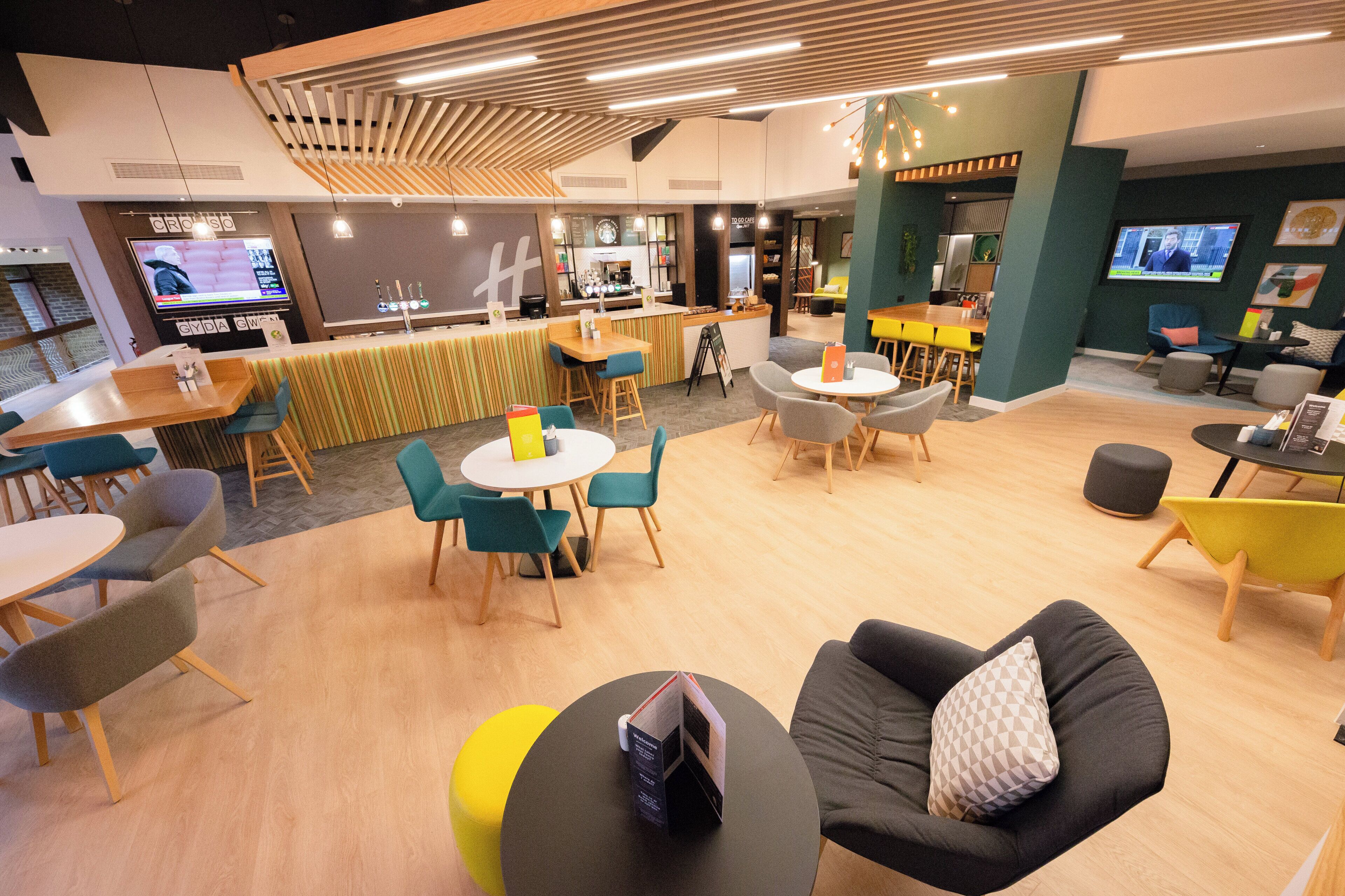 A modern lounge area with multiple seating options including sofas, chairs, and tables. There is a bar area with a counter and stools, and a television screen displaying a news program.