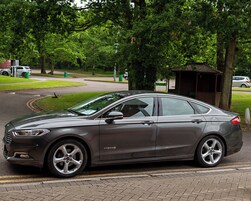 A dark-colored sedan parked on a paved area with a grassy field and trees in the background.