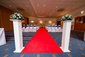 Corridoio di un hotel con tappeto rosso che conduce a un'area relax, pilastri bianchi con composizioni floreali e tappeto blu con motivo decorativo.