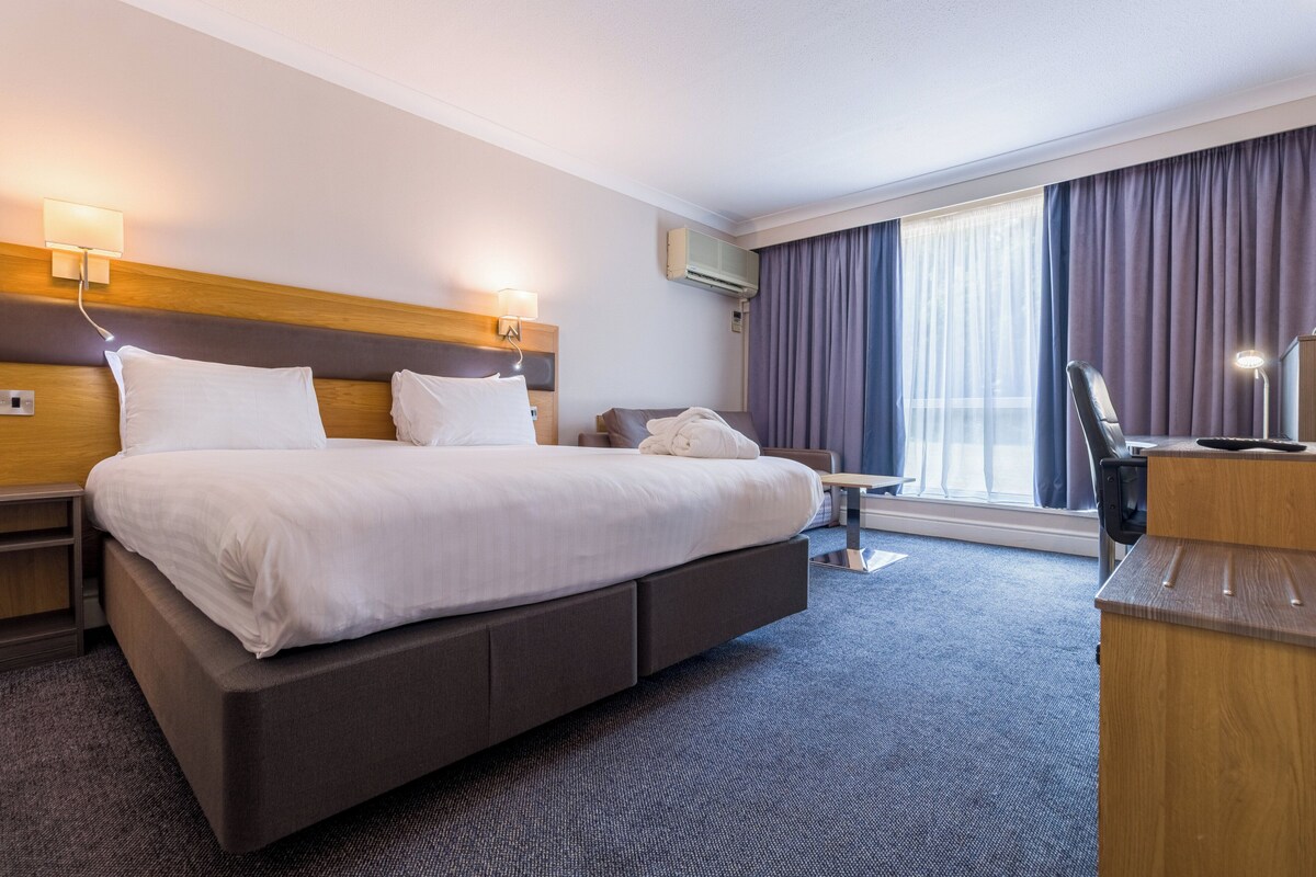 A hotel room with a large bed, bedside tables, a desk, and a window with curtains.