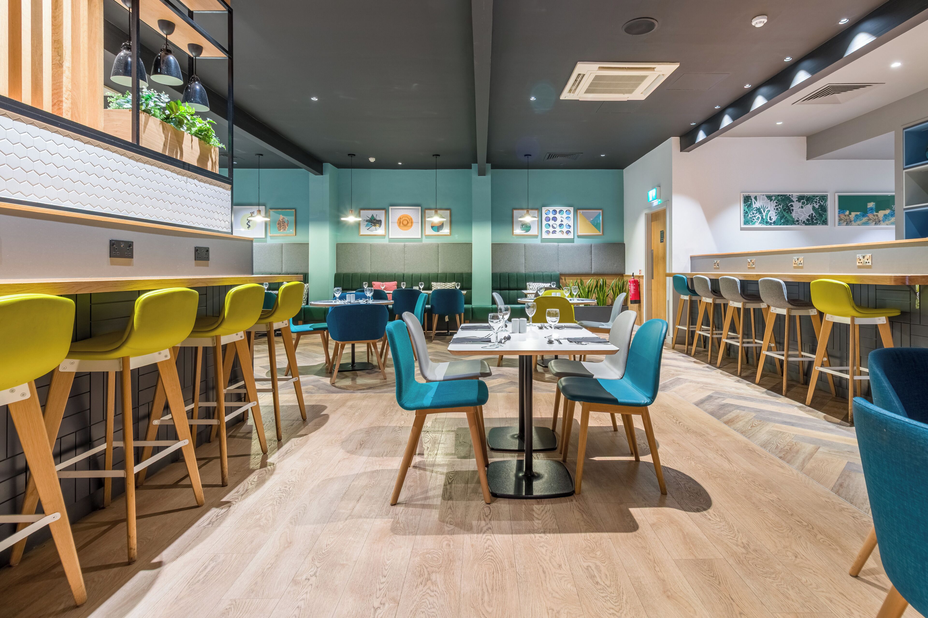 A modern restaurant interior with wooden bar counters, teal and yellow chairs, and a herringbone patterned floor.
