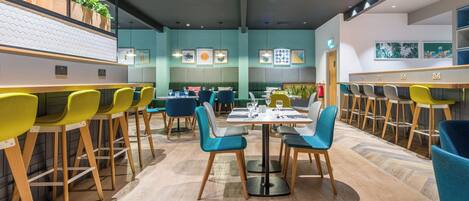 A modern restaurant interior with wooden bar counters, teal and yellow chairs, and a herringbone patterned floor.