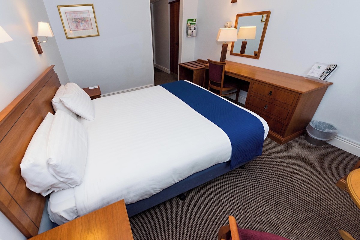 Una camera d'albergo con un letto di legno, una scrivania, una sedia, un comò e due lampade a parete.