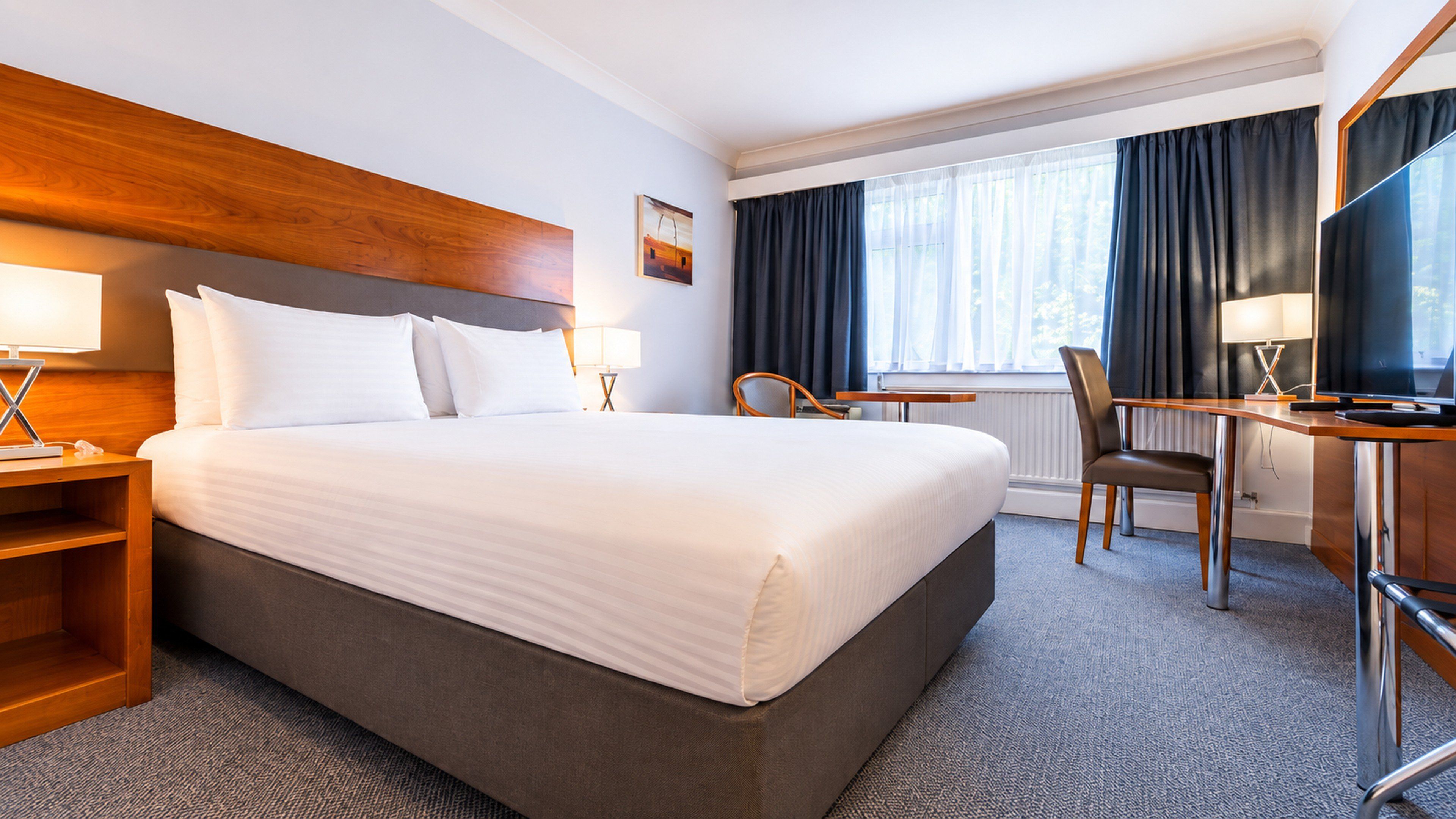 A hotel room with a large bed, a desk, a chair, a television, and a window with curtains.