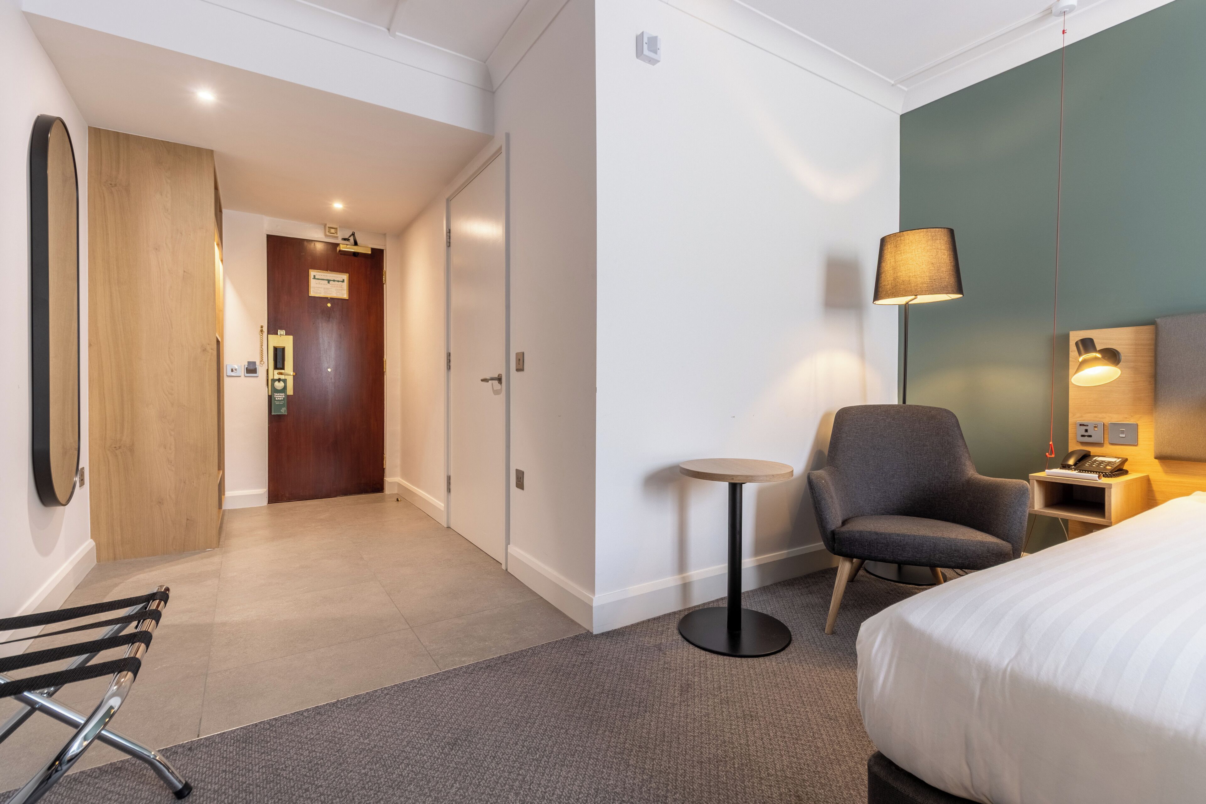 Ein Hotelzimmer mit einem Bett, einem Sessel, einem kleinen Tisch, einer Lampe und einem Schreibtisch mit Telefon.