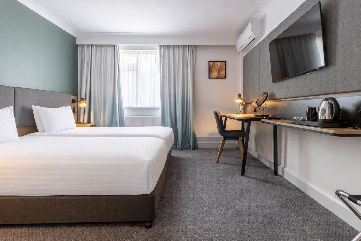 A hotel room with a bed, a desk, a chair, a flat-screen TV, and an air conditioning unit.