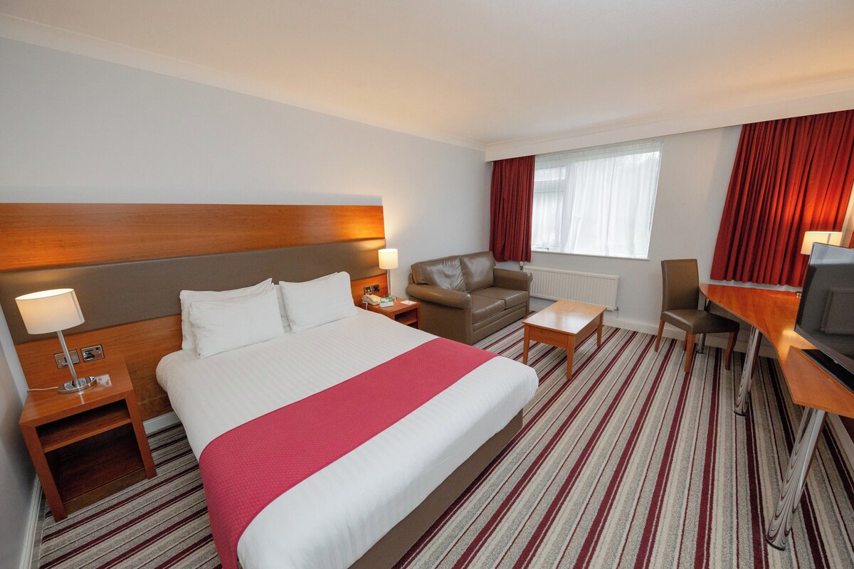 A hotel room with a large bed, a sofa, a desk with a chair, a TV, and a window with curtains.