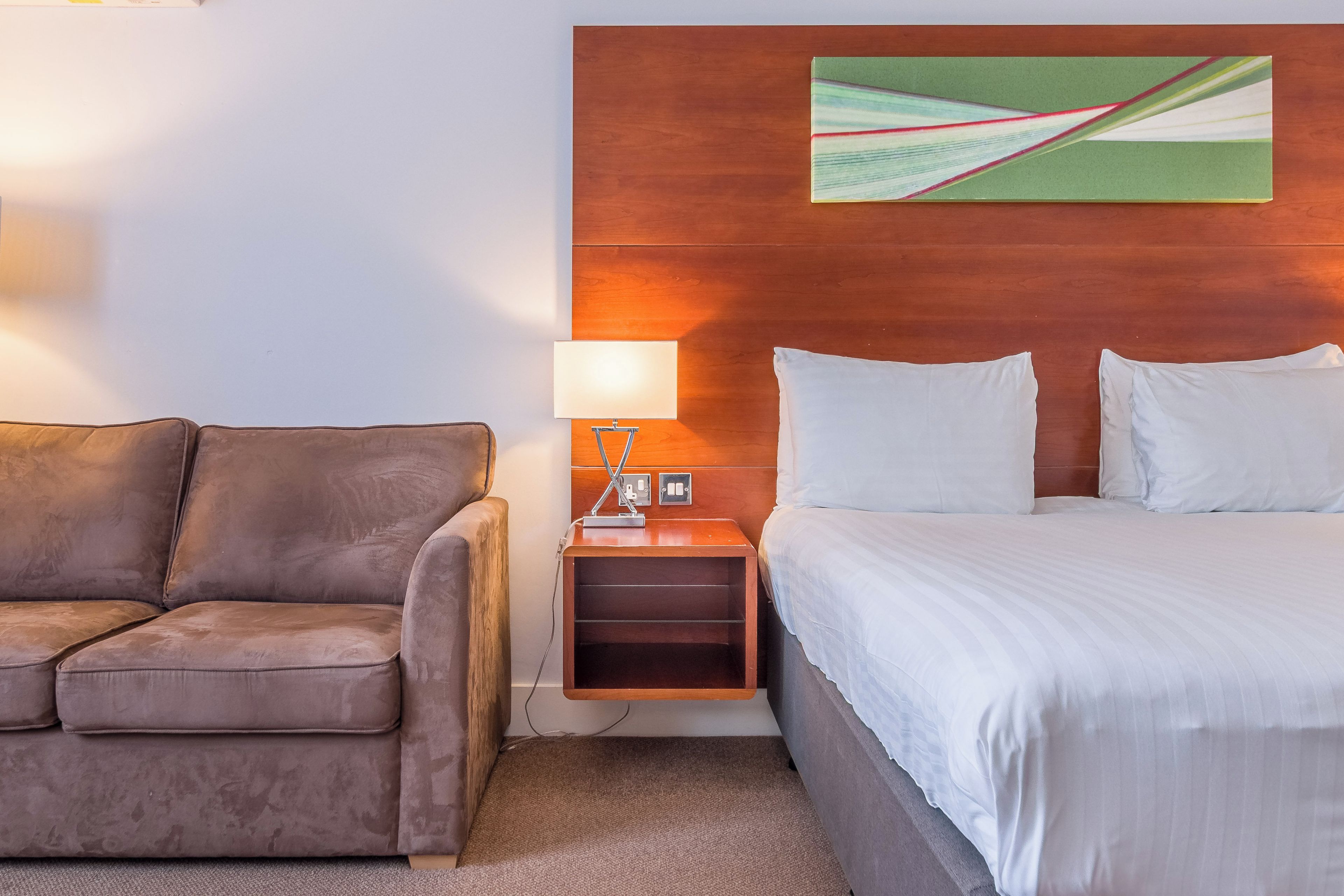 A hotel room with a bed, a nightstand, a lamp, and a sofa.