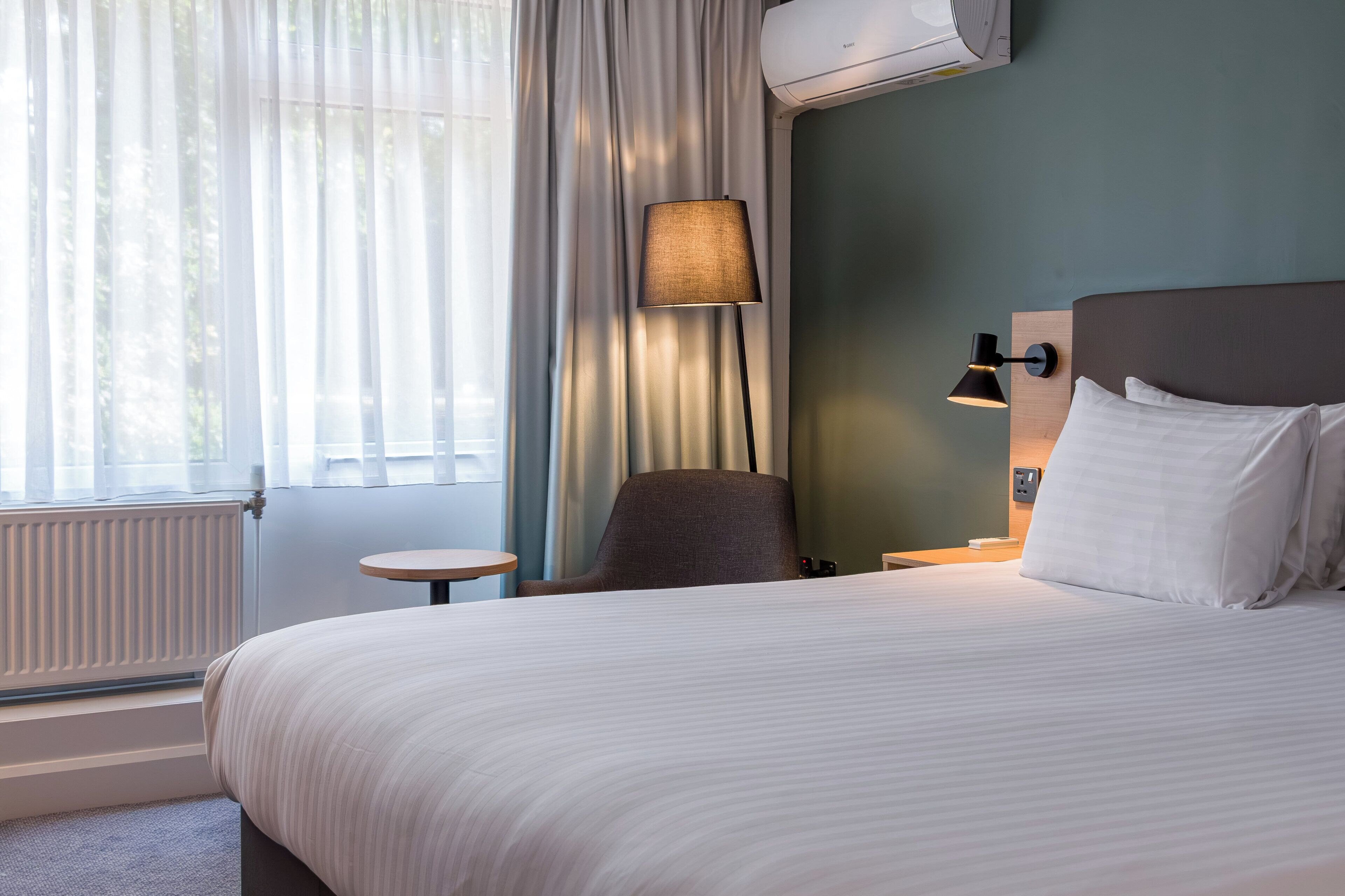 Une chambre d’hôtel équipée d’un lit, d’une chaise, d’une table, d’une lampe et d’un appareil de climatisation.