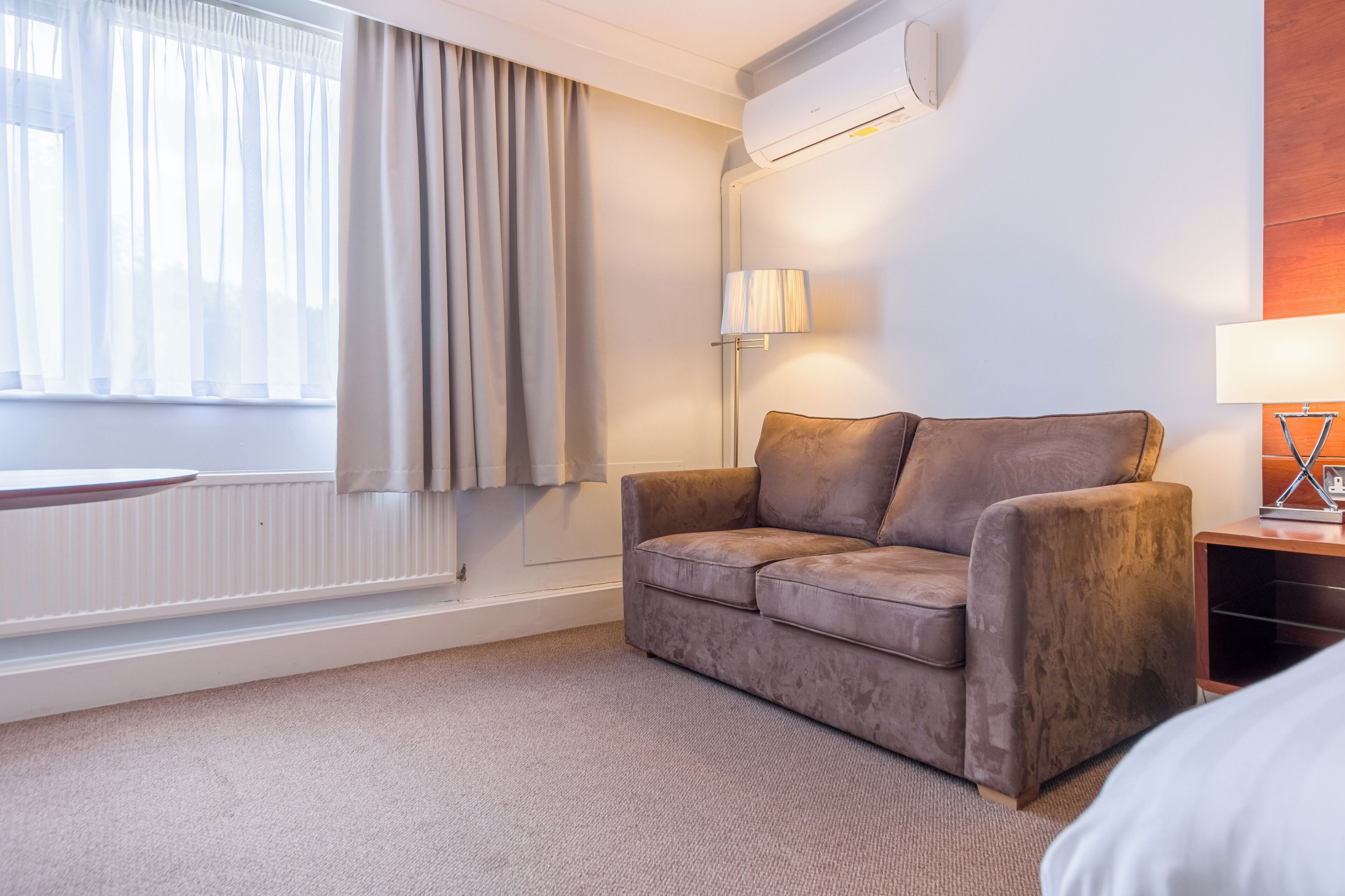 Ein Hotelzimmer mit einer Couch, einer Lampe, einem Heizkörper und einem Fenster mit Vorhängen.