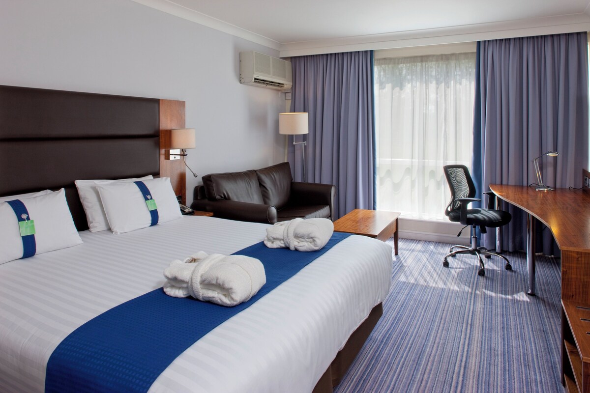 A hotel room with a large bed, a desk, a chair, a lamp, and a window with curtains.